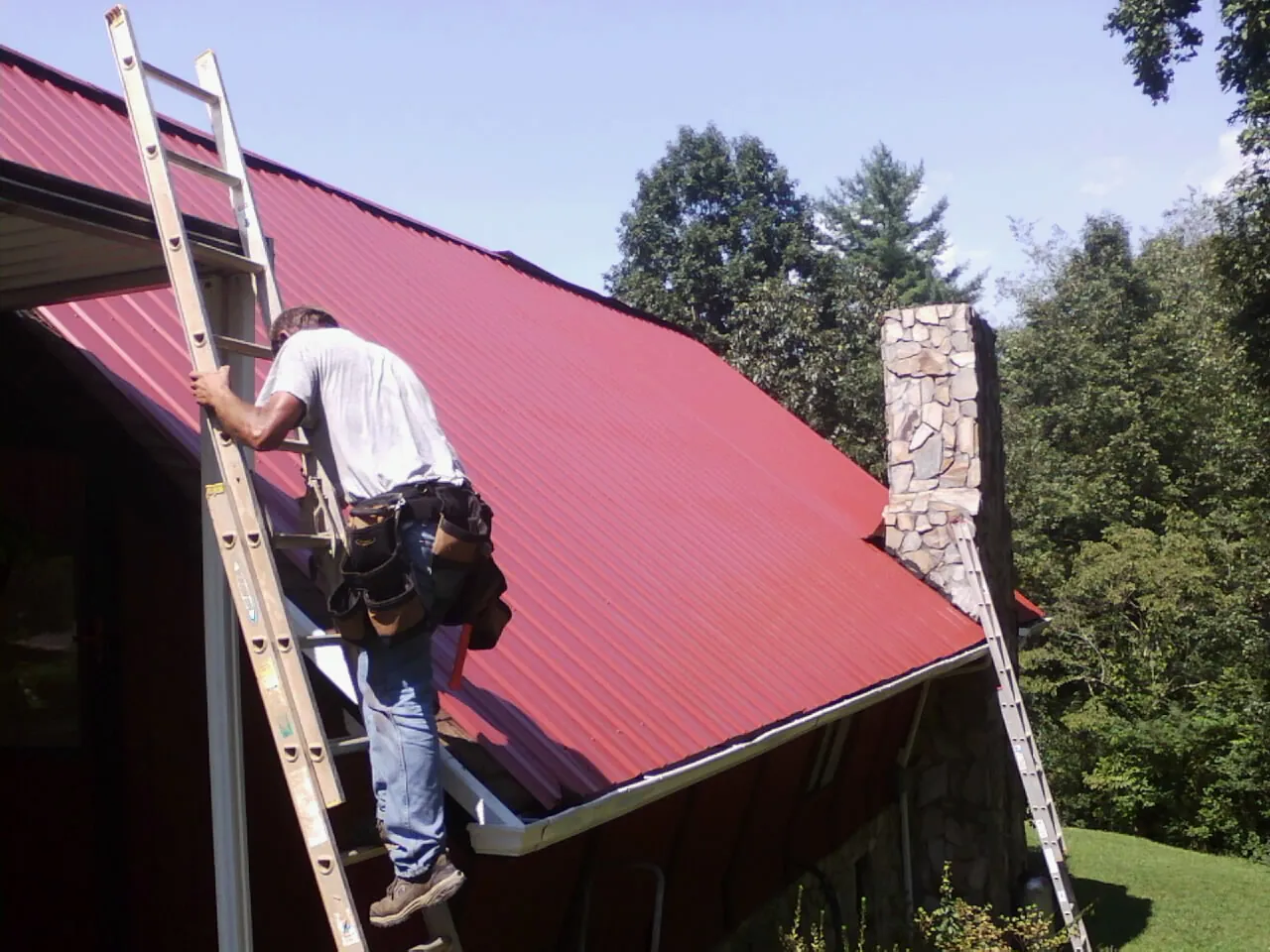 Roofer on a ladder beside a red metal roof and stone chimney — Soffit & Fascia Repair by Roofing Brothers in State Road